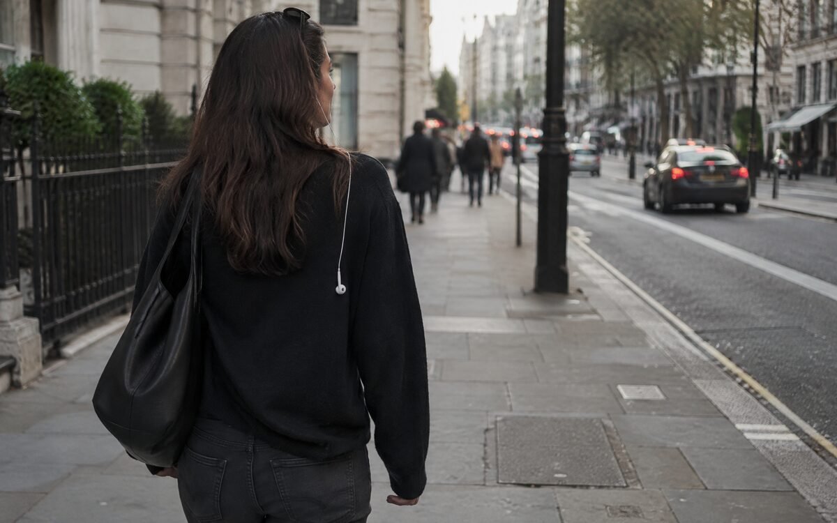 A person walking a city street with one earbud out, phone away, head up