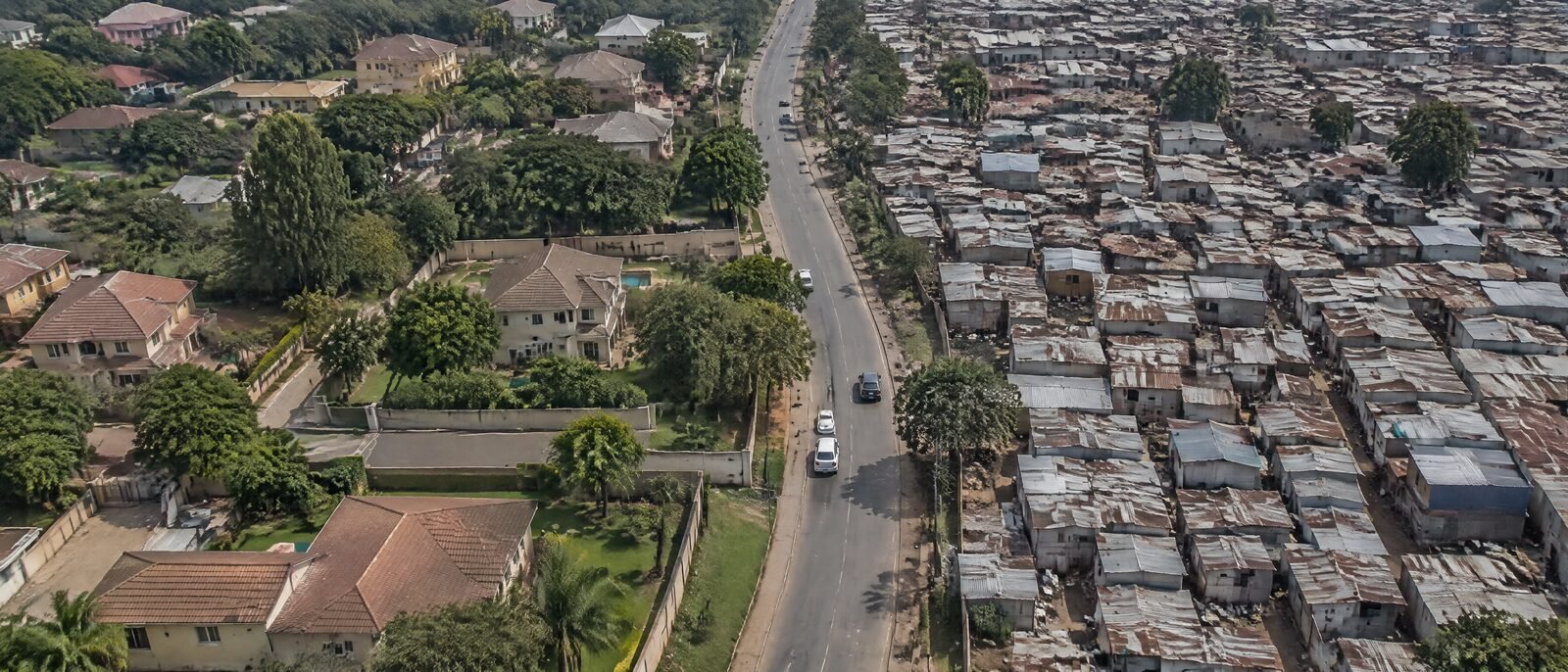 A split view — a calm leafy suburban street on one side, a dense informal settlement on the other, same city