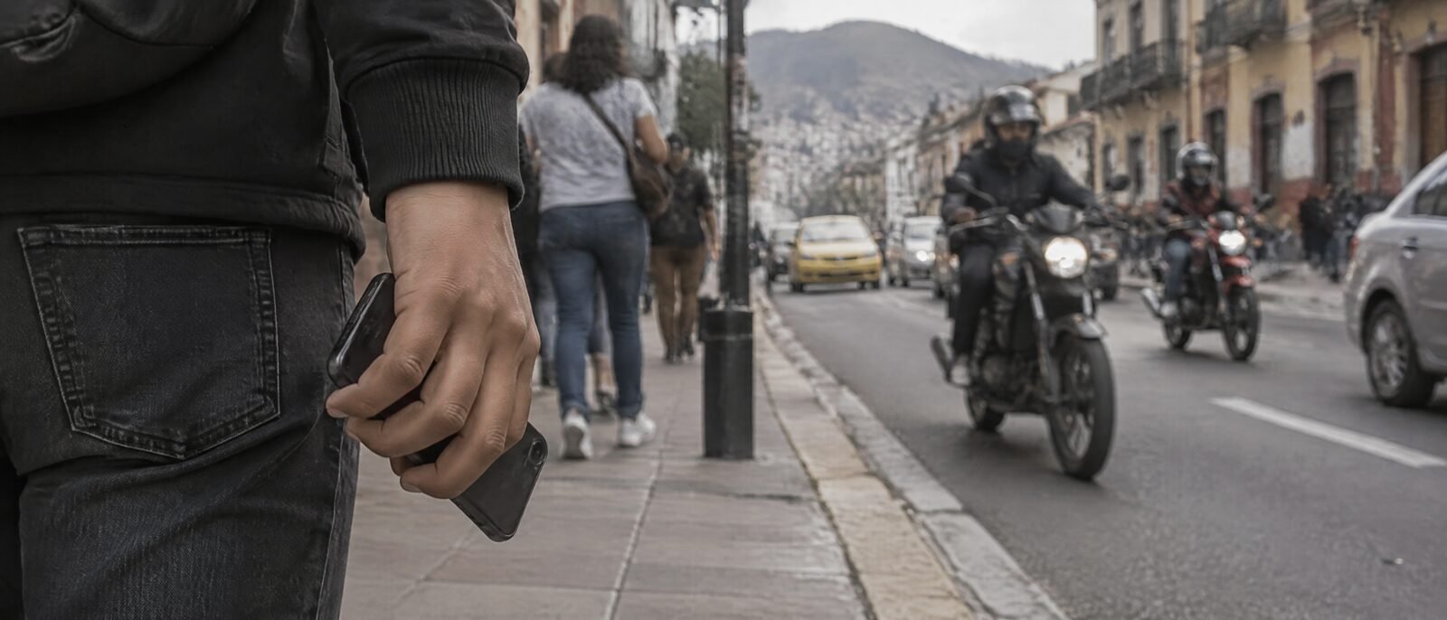 A hand holding a phone low and close to the body on a busy Colombian city pavement, motorbikes passing in the road behind