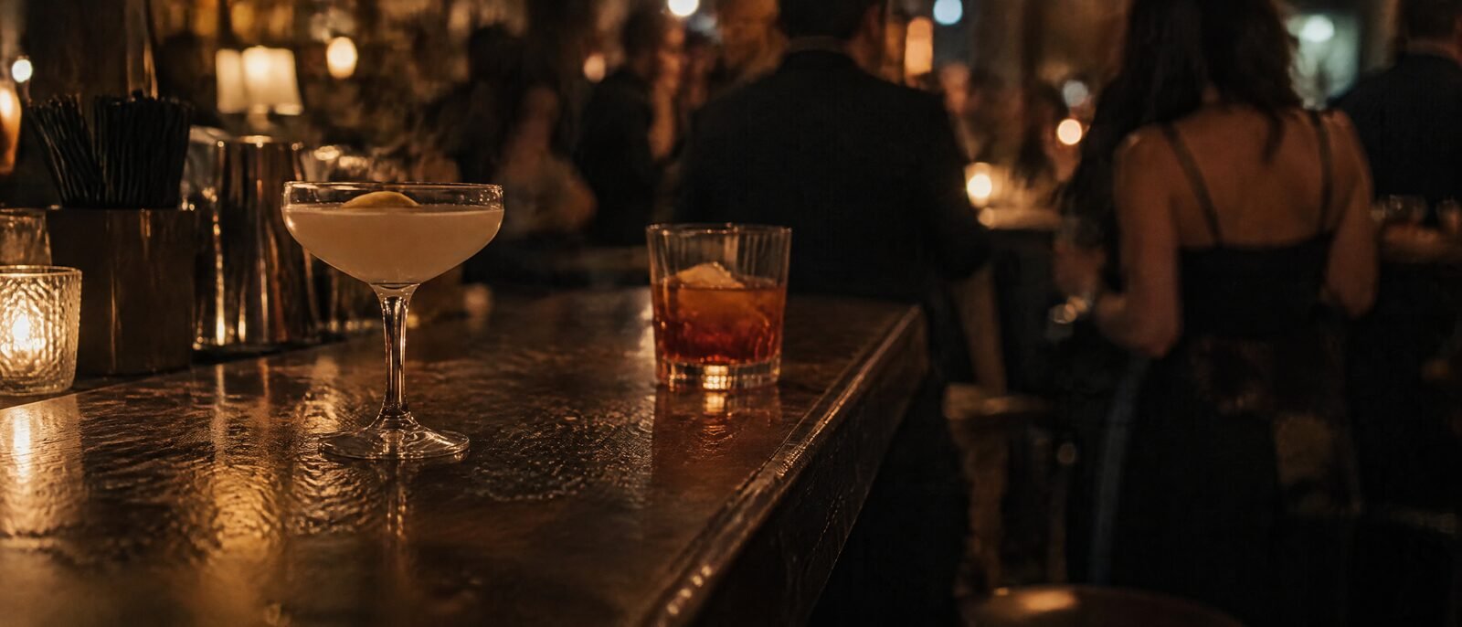 Two cocktail glasses on a dark bar counter under warm low light, one unattended, nightlife blurred behind