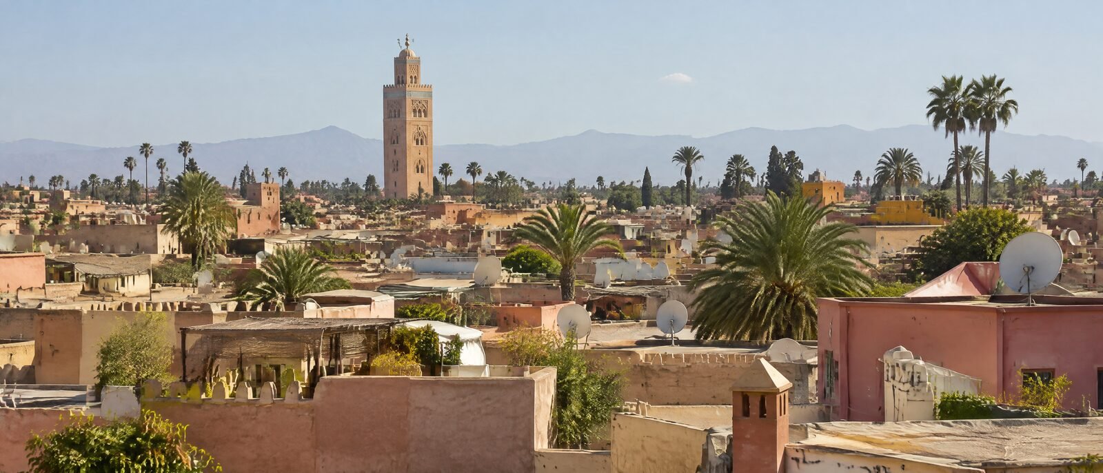 A wide daylight view over the rooftops of Marrakech medina toward the Koutoubia minaret, terracotta buildings, clear sky
