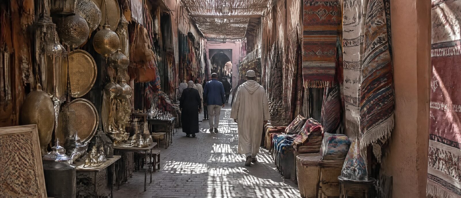 A narrow Marrakech medina alley with market stalls on both sides, a few people walking, daylight